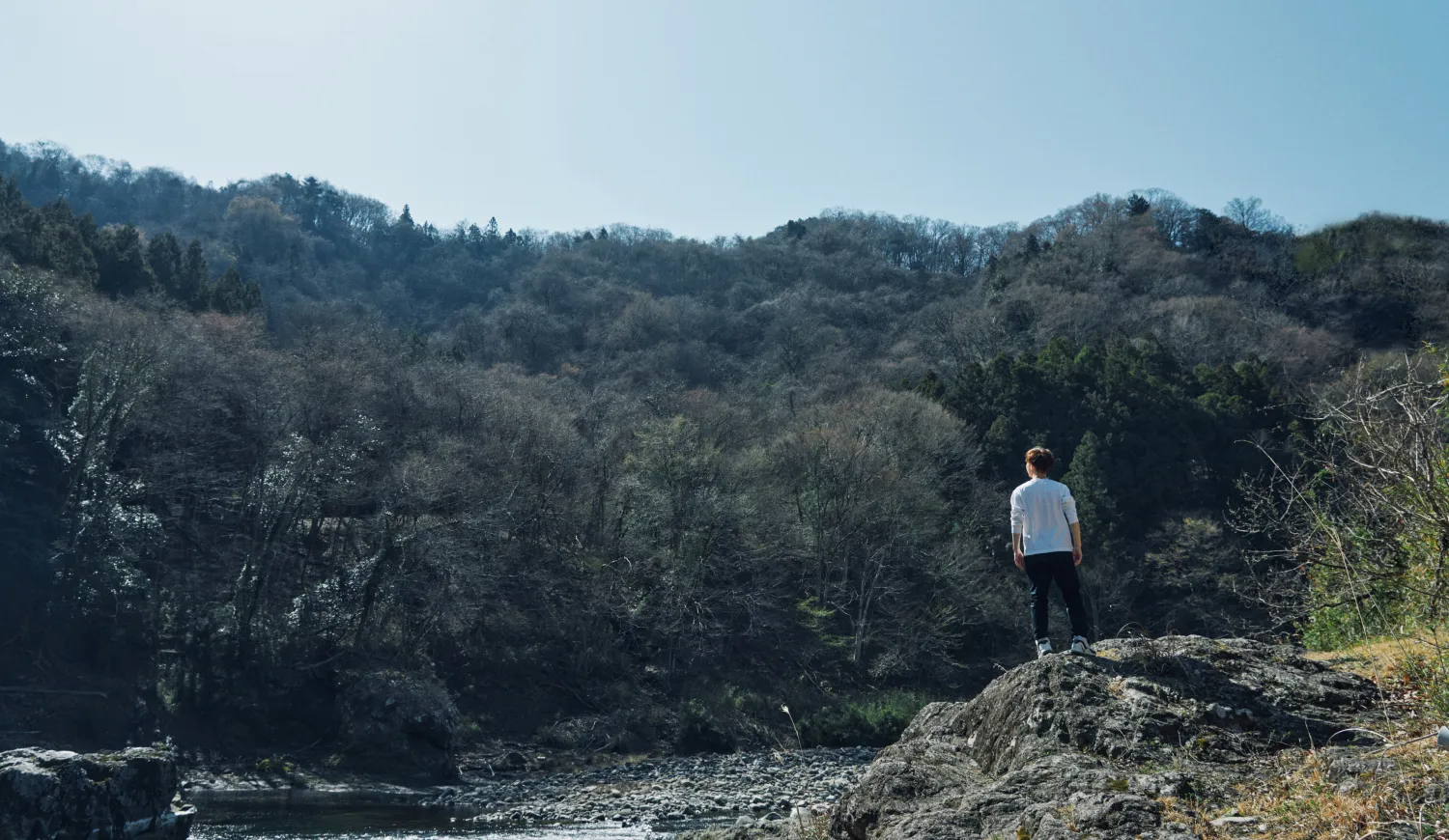 川辺の岩の上に立ち、山あいの景色を見つめる人物